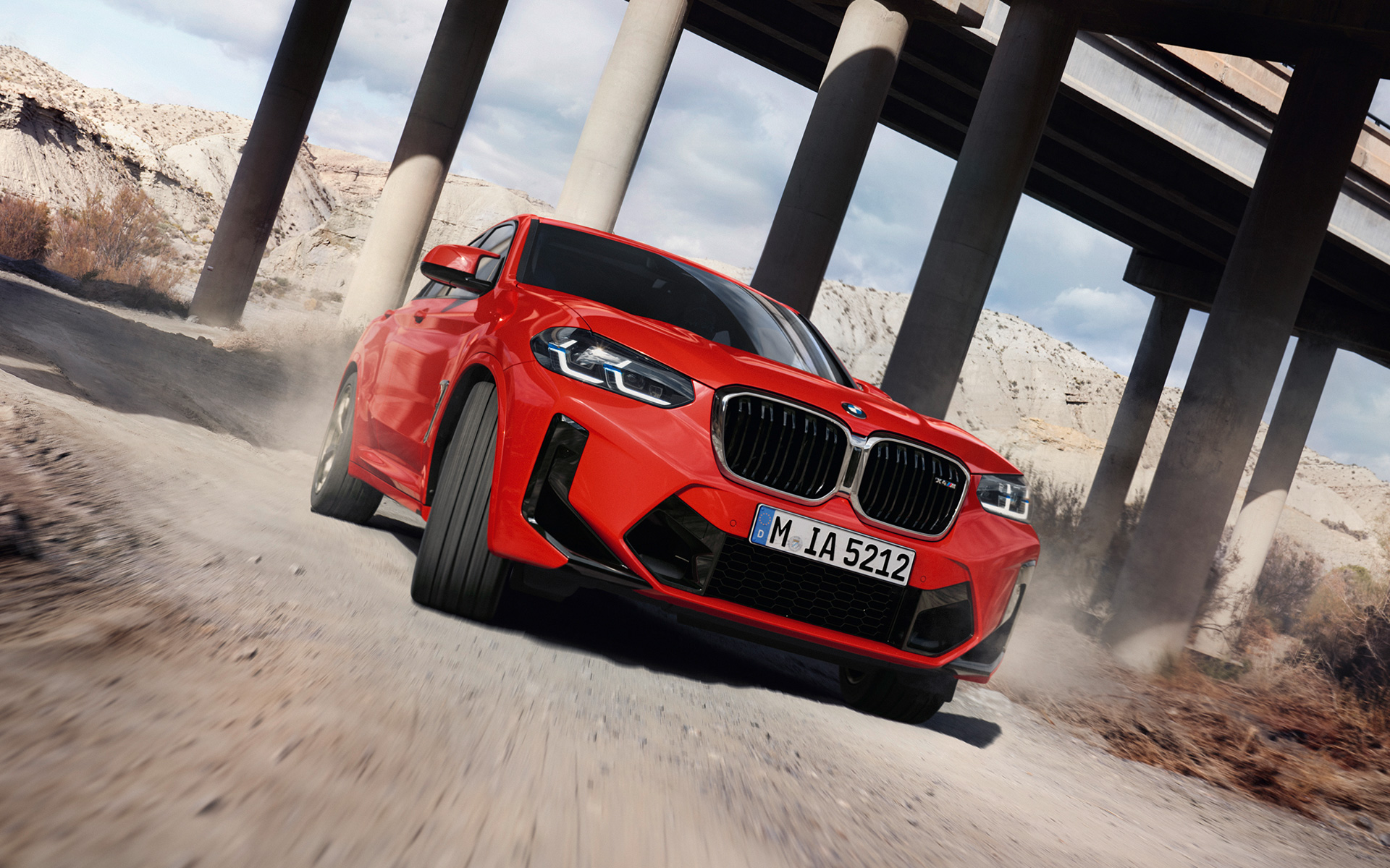 مشهد جانبي من الزاوية الأمامية للسيارة BMW X4 M F98 LCI Facelift 2021 أمام أعمدة الجسر بلون أحمر معدني تورنتو  